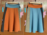 Two A-line skirts from BUTIK KUBIK hang against a brown background. On the left is the Kubik skirt Twist short waffle multicolour size S with orange, blue, and dark grey panels; the right features light blue with orange, olive, and grey accents.