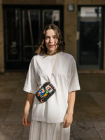 A person with shoulder-length hair stands outside in a loose white outfit, carrying the YKRA travel MAGIC SEALIFE bag by BUTIK KUBIK, featuring an adjustable cotton strap. The background is blurred, revealing dark glass doors and a tiled floor.