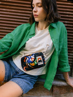 A person in a green shirt over a white t-shirt and denim shorts leans against a corrugated wall, gazing thoughtfully. They have a colorful patterned bag with travel essentials like the BUTIK KUBIK YKRA travel MAGIC SEALIFE featuring a vintage-style YKK zipper.