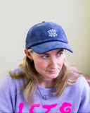 A woman with long blonde hair wears a BUTIK KUBIK Tropical Scandinavia corduroy cap in blue and a lavender sweatshirt with pink letters, looking slightly to her left against a plain, light-colored background.