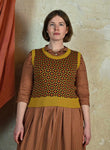 A woman with short brown hair stands against a beige wall, wearing a patterned brown dress and the Palava Shirley - Mustard Patchwork Vest from BUTIK KUBIK, made of organic cotton yarn. An orange curtain is visible to her left.
