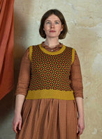 A woman with short brown hair stands against a beige wall, wearing a patterned brown dress and the Palava Shirley - Mustard Patchwork Vest from BUTIK KUBIK, made of organic cotton yarn. An orange curtain is visible to her left.