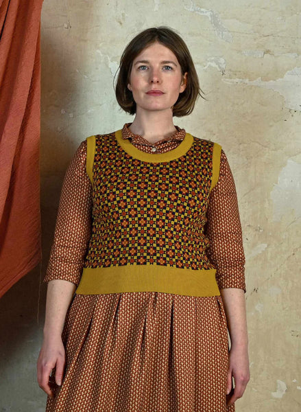 A woman with short brown hair stands against a beige wall, wearing a patterned brown dress and the Palava Shirley - Mustard Patchwork Vest from BUTIK KUBIK, made of organic cotton yarn. An orange curtain is visible to her left.