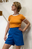 A person wearing a fitted orange t-shirt with white trim and high-waisted Kubik workwear shorts in vintage fabric by BUTIK KUBIK stands in front of a white wall, looking to the side with one hand on their hip.