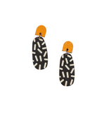 The Törmi Neilikka oval earrings by BUTIK KUBIK feature an orange semi-circular top and a black oval bottom adorned with white sprinkle-like patterns. Made from Finnish birch plywood, each pair is packaged sustainably to enhance your eco-friendly style.