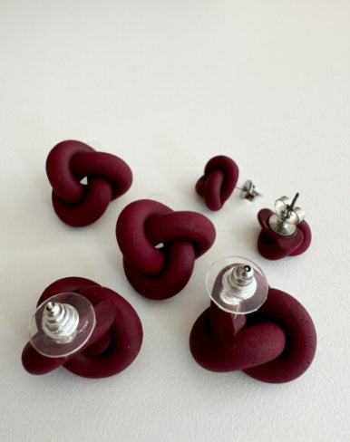 Five pairs of Ochform örhängen knut stor by BUTIK KUBIK, made of burgundy clay and shaped into stylish knots, displayed on a white surface with clear plastic backings and silver posts.