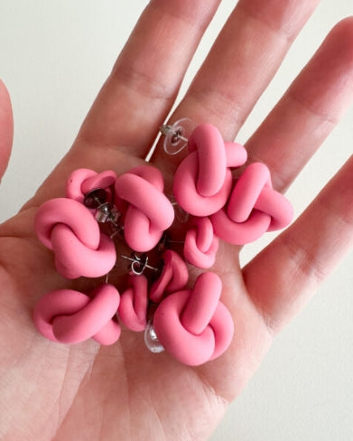 A hand holds several pairs of Ochform örhängen knut liten by BUTIK KUBIK—playful, matte, pink polymer clay stud earrings with a twisted design and clear plastic backings.