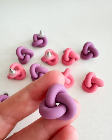 A hand holds a purple Ochform örhängen knut stor by BUTIK KUBIK, made of clay. In the background, several similar pink and purple handmade earrings are scattered on a white surface.