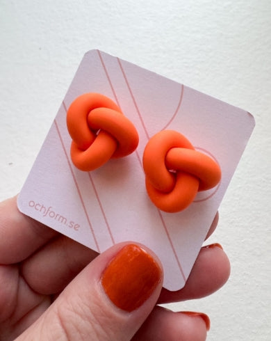 A hand with orange-painted nails holds a card from BUTIK KUBIK displaying Ochform örhängen knut liten—unique, bright orange polymer clay knot earrings—featuring a minimal line design and ochform.se in the corner.