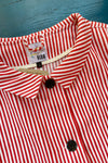 A close-up of the Kubik Overshirt Strömma Kadett rödvit by BUTIK KUBIK, featuring red and white vertical stripes, collar, black buttons, and a KUBIK size S label. Made in Sweden from 100% cotton vintage deadstock fabric on a blue surface.