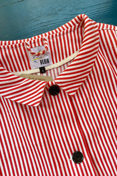 A close-up of the Kubik Overshirt Strömma Kadett rödvit by BUTIK KUBIK, featuring red and white vertical stripes, collar, black buttons, and a KUBIK size S label. Made in Sweden from 100% cotton vintage deadstock fabric on a blue surface.