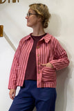 A person wearing glasses, a maroon shirt, blue pants, and the BUTIK KUBIK Kubik cotton shirt striped with large pockets stands against a plain white wall, looking to the side.