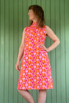 A woman stands against a green paneled wall, wearing the sleeveless, high-neck Kubik dress cineast Popsicle by BUTIK KUBIK with a bold pink and white circular pattern, looking to the side with one hand on her hip.