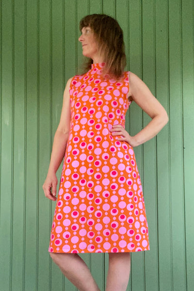 A woman stands against a green paneled wall, wearing the sleeveless, high-neck Kubik dress cineast Popsicle by BUTIK KUBIK with a bold pink and white circular pattern, looking to the side with one hand on her hip.