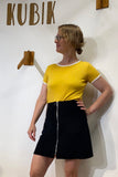 A woman wearing glasses and the Kubik 70s T-shirt in yellow by BUTIK KUBIK, paired with a black skirt, stands in front of a white wall with "KUBIK" written on it. She looks to the side with one hand on her hip.