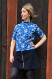 A woman stands before a dark wooden wall in the Kubik t-shirt Blåsippa by BUTIK KUBIK, a 60’s-inspired blue floral knit top, paired with a dark corduroy skirt with a front zipper. She poses confidently, one hand on her hip, gazing to the side.