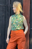 A person with light hair stands in front of a dark wooden wall, wearing the BUTIK KUBIK Kubik top cineast Baldersbrå—a sleeveless green floral organic cotton top—and high-waisted orange pants, with one hand in their pocket and looking to the side.