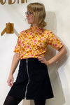 A person with blonde hair and glasses poses against a white wall wearing the BUTIK KUBIK "Kubik t-shirt 1966," a 60's inspired knit floral top, paired with a black skirt featuring a front zipper and large pockets.