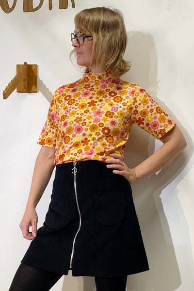 A person with blonde hair and glasses poses against a white wall wearing the BUTIK KUBIK "Kubik t-shirt 1966," a 60's inspired knit floral top, paired with a black skirt featuring a front zipper and large pockets.