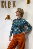 A woman in glasses wears the Kubik sweater mushroom by BUTIK KUBIK—a 60s-inspired knit top made in Sweden—paired with orange pants, and stands in front of a white wall with KUBIK and wooden decor, looking to the side with one hand on her hip.