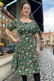 A woman with tattoos and dark hair tied back smiles on a cobblestone street, wearing the BUTIK KUBIK Kubik dress Twinkle dress Midi Baldersbrå—a green floral 50s-inspired short-sleeve trikåklänning with black tights. Buildings and parked cars are behind her.