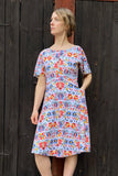 A woman stands before a dark wooden wall, wearing the BUTIK KUBIK Kubik dress Twinkle Midi Dalarö with a colorful floral and star print in red, blue, and yellow. She gazes calmly to the side with a serene expression.