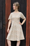 A woman in the Kubik dress Twinkle Midi Linblom by BUTIK KUBIK, a short-sleeved white floral dress with a brown pattern, stands by a rustic wooden door, looking aside with one hand on her hip.