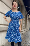 A woman smiles on a cobblestone street, wearing BUTIK KUBIK’s Kubik dress Twinkle dress Midi Blåsippa—en blommig blå klänning med vitmönster—plus svarta strumpbyxor och visar tatueringar på armarna.