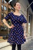 A woman with tattoos stands on a city sidewalk, wearing the BUTIK KUBIK Kubik dress Twinkle dress Mini Nattviol, which features a colorful flower pattern. She smiles slightly upward with buildings and a shop window behind her.