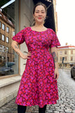 A person stands on a cobblestone street wearing the vibrant pink and red Kubik dress Twinkle dress Midi Nejlika by BUTIK KUBIK, smiling with visible arm tattoos. Buildings and parked cars are in the background.