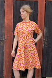 A woman poses by a rustic wooden door, wearing the Kubik dress Twinkle crew neck retro 1966 from BUTIK KUBIK, featuring a colorful orange, yellow, and pink floral pattern. She looks to the side with her hand on her hip.