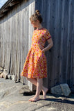 A woman wearing the BUTIK KUBIK Kubik dress Twist 1966, made from organic GOTS fabric, stands barefoot on rocks beside a weathered wooden building, looking down and to the side with her hands in her pockets.