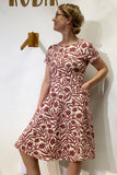 A woman in glasses wears the Kubik dress Twist Botan Burgundy by BUTIK KUBIK—a short-sleeved, knee-length dress featuring a red and white botanical pattern, crafted from ökotex-certifierat sweatshirttyg—standing sideways with one hand in her pocket against a white wall.