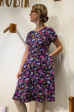 Indoors, a person with tied-back hair and glasses wears the Kubik dress Twist Rosemary by BUTIK KUBIK—a 50s-inspired, short-sleeved, knee-length floral cotton jersey dress—with one hand in the pocket, looking to the side.