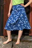 A person stands barefoot on rocks wearing the Kubik skirt Twist Midi Blåsippa from BUTIK KUBIK, styled with a light green polka dot top. Behind them is a wooden wall with dark and reddish-brown panels.