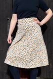A person in a black short-sleeve top and the Kubik skirt Twist Linblom by BUTIK KUBIK—an eco-friendly cotton linen floral midi with a small brown pattern—stands before a dark wooden door, hand on hip.