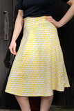 A person stands with one hand on their hip, wearing a black top and the BUTIK KUBIK Kubik skirt Twist Midi Trio—yellow, 50s-inspired, and patterned with small white triangles—in front of a dark wooden door.