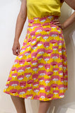 A woman wearing a yellow top and the BUTIK KUBIK "Kubik skirt Twist Midi Water Lily," a 50s-inspired, knee-length jersey skirt with a pink, yellow, and white floral pattern, stands against a white wall with one hand on her hip.