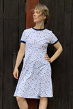 A person stands before a dark wooden wall, wearing the BUTIK KUBIK Kubik dress Twist Strandfynd—a 50s-inspired OEKOTEX-certified cotton dress featuring a colorful confetti pattern and navy trim. They look to the side with one hand on their hip.