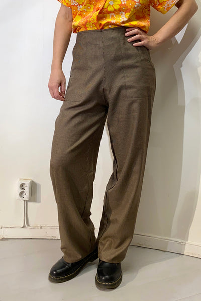 A person stands indoors near a white wall and outlet, wearing BUTIK KUBIK's Kubik trousers wool silk in brown, a bright orange floral shirt, and black shoes. Their head is not visible in the image.