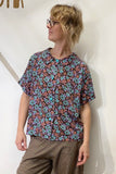 A person wearing the BUTIK KUBIK Kubik blouse vintage, an oversized boxy short-sleeve button-up with a blue and brown floral pattern, paired with brown pants, stands against a plain light-colored wall looking to the side.