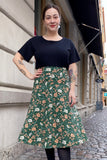 A woman with tied-back dark hair, tattoos, and earrings stands on a cobblestone sidewalk wearing a black top and the BUTIK KUBIK Kubik skirt Twist Midi Baldersbrå with green floral prints. Beige buildings and shop awnings are in the background.