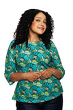 A person with curly dark hair, wearing the Cissi och Selma top Vendela Kronblad Turkos by BUTIK KUBIK—featuring turquoise, green, and yellow floral patterns—and dark jeans, stands against a white background, smiling gently with hands clasped.