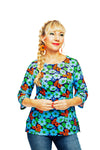 A smiling person with long braided blonde hair wears the vibrant Cissi och Selma top Vendela humlesurr blå from BUTIK KUBIK, featuring blue, orange, and green patterns on ekologisk bomullstrikå. They also have red earrings and are set against a white background.