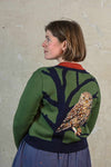 A woman with chin-length brown hair stands sideways, wearing the BUTIK KUBIK Palava Vera green owl cardigan, which features a jacquard knit with a large tree branch and detailed owl design on the back, in front of a textured, light-colored wall.