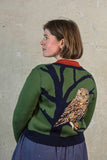 A woman with chin-length brown hair stands sideways, wearing the BUTIK KUBIK Palava Vera green owl cardigan, which features a jacquard knit with a large tree branch and detailed owl design on the back, in front of a textured, light-colored wall.