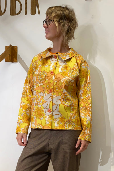 A person with short, wavy blonde hair and glasses stands against a white wall wearing brown pants and the colorful Kubik Overshirt retro by BUTIK KUBIK, featuring a yellow and orange floral cotton deadstock fabric.