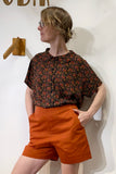 Wearing glasses, the person stands with hands in pockets and looks to the side against a plain wall. They wear high-waisted orange shorts and a BUTIK KUBIK Kubik blouse floral viscose.