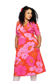 A woman with curly hair wears the BUTIK KUBIK Cissi och Selma Vivi dress 1960 Rosa, a bright orange and pink floral cotton jersey dress with short sleeves and v-neck. She stands against a white background, smiling and looking to the side.