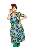 A cheerful woman poses playfully in a vibrant Cissi och Selma Vivi humlesurr blå dress by BUTIK KUBIK, featuring blue, green, and red patterns. Complemented by green tights, she has one hand to her ear and the other on her hip, with hair styled up and wearing dangling earrings.